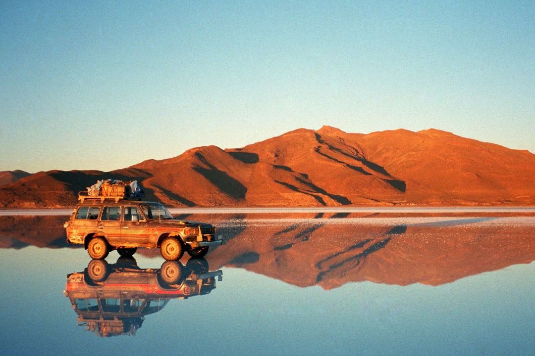 imagen de un vehiculo en el salar de uyuni, en temporada de lluvia de fondo una montaña y con el reflejo espejo tradicional del salar de uyuni.